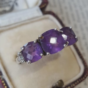May include: A silver ring featuring three cushion-cut purple amethyst gemstones set in a row. The band is made of polished silver and includes small decorative metal accents on the sides near the base of the primary purple stone settings.
