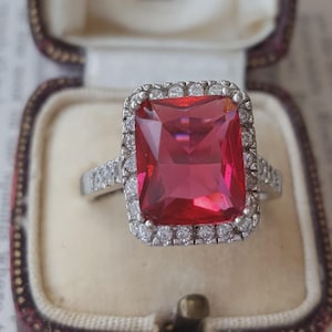 Op de afbeelding: Een zilverkleurige ring met een grote, rechthoekige, felrode middensteen. De centrale edelsteen is omringd door een halo van kleine, heldere, ronde steentjes, met extra heldere steentjes gezet langs de band van de ring.