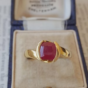 Può includere: Un anello in oro con un rubino rosso centrale a taglio cuscino. La pietra è trattenuta da una montatura in oro liscio e lucido che avvolge i lati. Il gambo è in oro massiccio con un design affusolato, presentato in un cofanetto blu.