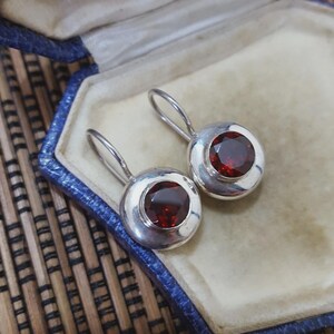 May include: Silver drop earrings featuring large, round, deep red gemstones. The earrings are set in a polished silver bezel. They are presented in a blue jewellery box with a cream interior.