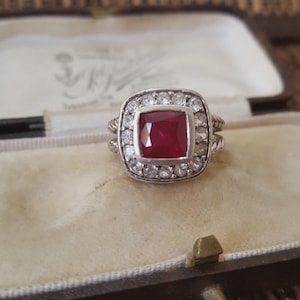 May include: A silver ring featuring a square, crimson gemstone at its centre, encircled by a halo of smaller, clear stones. The ring is set in a vintage style, presented in a velvet-lined jewellery box. The ring's band has a twisted rope design.
