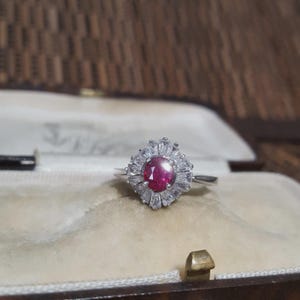 Op de afbeelding: Een zilveren ring met een ovale robijn in het midden, omringd door kleinere, heldere edelstenen. De ring heeft een vintage design en wordt gepresenteerd in een bruine sieradendoos. De robijn is dieprood.