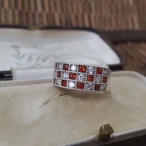 May include: A silver ring with a rectangular design, featuring alternating rows of red and clear gemstones. The ring is displayed inside a vintage jewellery box with a cream-coloured interior.