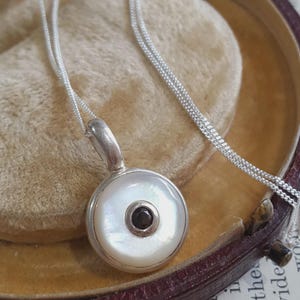 Peut inclure: Un collier en argent avec un pendentif circulaire incrusté de nacre blanche et une petite pierre précieuse ronde d'un noir profond au centre. Le pendentif est suspendu à une fine chaîne en argent, présentant une finition métallique polie.