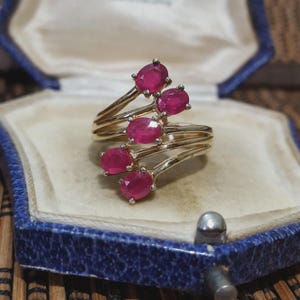 May include: A gold ring set with five oval, vivid red gemstones. The ring has a swirling, layered design, with each gemstone held by prongs. The ring is presented inside a blue jewellery box.