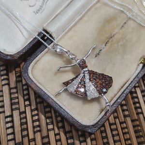 Può includere: Collana con pendente a ballerina in argento. Il pendente raffigura una ballerina con un abito di pietre preziose marrone e bianco. La collana è esposta in un portagioie vintage con interno color crema ed esterno marrone scuro.