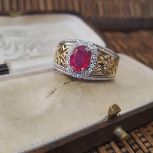 May include: A two-tone ring featuring a large oval ruby gemstone, encircled by small clear stones. The ring showcases a gold filigree pattern on the sides and a silver band. It's presented in a vintage jewellery box.