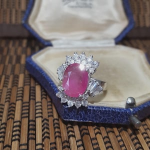 Op de afbeelding: Zilveren ring met een grote ovale robijn, omringd door kleinere heldere edelstenen. De ring is gezet in een zilveren band en wordt gepresenteerd in een blauwe sieradendoos.