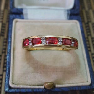 May include: A gold band ring featuring a row of four oval-cut red gemstones alternating with small, clear, round-cut accent stones. The ring is set in a polished yellow gold metal band, showcasing a classic channel-style setting design.
