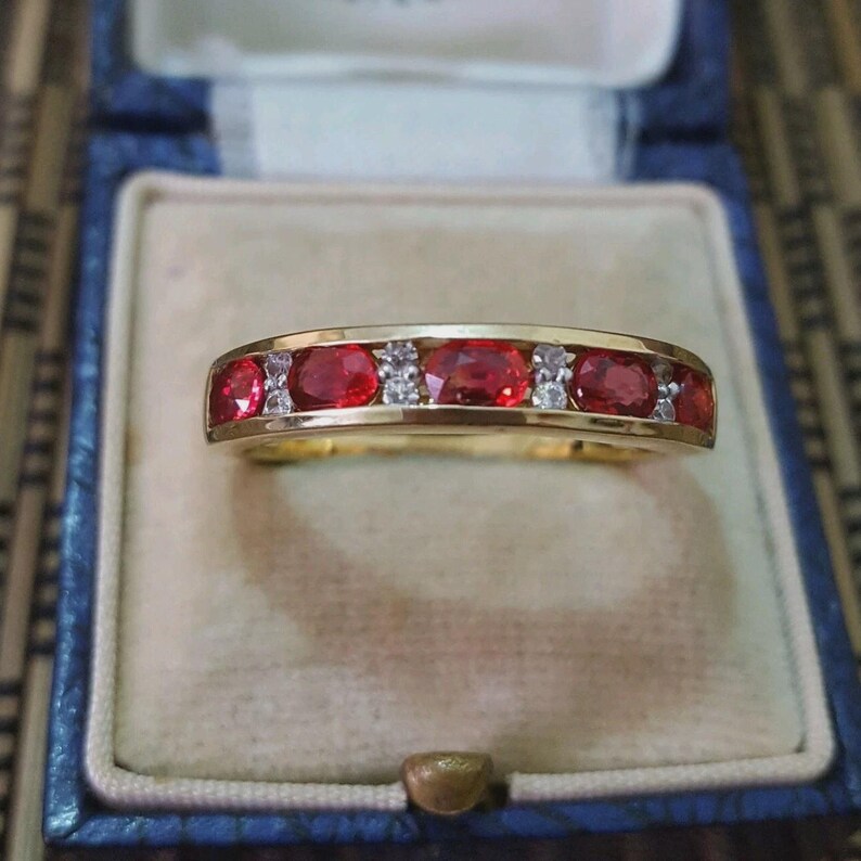 May include: A gold band ring featuring a row of four oval-cut red gemstones alternating with small, clear, round-cut accent stones. The ring is set in a polished yellow gold metal band, showcasing a classic channel-style setting design.