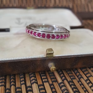 Könnte beinhalten: Ein silberner Ring mit einer Reihe kleiner, runder, leuchtend rosa Edelsteine. Der Ring befindet sich in einer Schmuckschatulle im Vintage-Stil mit hellem Innenraum und dunkelbraunem Äußeren. Die Schatulle liegt auf einer Bambusmatte.