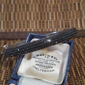 Könnte beinhalten: Ein silbernes Armreif-Armband mit schwarzen Edelsteinen, präsentiert in einer blauen Schmuckschatulle. Die Schatulle hat ein weißes Satinfutter und den Aufdruck "MARTIN & Co". Ein dunkles, elegantes Armband.