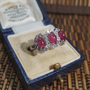May include: A silver ring featuring three oval, crimson gemstones, each encircled by smaller, clear stones. The ring is presented in a vintage blue jewellery box with a cream interior.