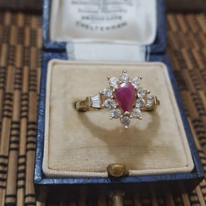 May include: A gold ring featuring a pear-cut ruby, encircled by a halo of clear gemstones. The ring is presented in a vintage blue jewellery box.