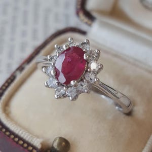Peut inclure: Une bague argentée ornée d'un rubis rouge foncé central de taille ovale, entouré d'une auréole de douze petites pierres blanches rondes et transparentes. La bague possède un anneau en métal poli et est présentée dans un écrin à bijoux.