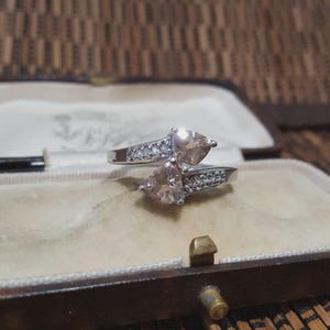 May include: A silver ring featuring two heart-shaped, peach-coloured gemstones. The band is decorated with small, clear stones. The ring is presented in an open, vintage jewellery box with a beige interior.