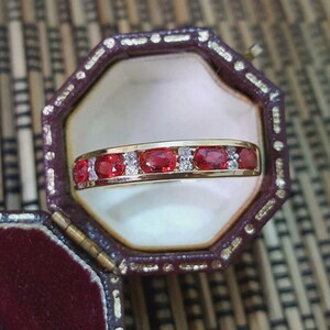 May include: A gold band ring featuring a row of alternating oval-cut red gemstones and small clear round stones, presented inside an octagonal dark red velvet-lined jewelry box with a gold-toned trim along the edges of the lid and base.