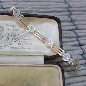 Könnte beinhalten: Ein silbernes Armband mit einer Reihe von klaren Zirkonia-Steinen. Das Armband befindet sich in einer samtenen Box mit einem Etikett, auf dem steht "Makers to the Admiralty. R. Gilbert & Sons. Goldsmiths & Jewellers. 12 Broadgate & 21 Burgess Coventry. Also at Barrow & Dalton."