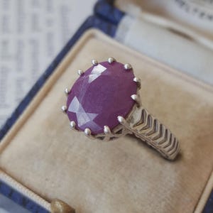 Peut inclure: Une bague en argent ornée d'une grande pierre précieuse ovale de couleur magenta profond, maintenue par un sertissage à griffes. L'anneau présente un motif texturé en forme de chevrons sur sa surface extérieure, partant du sertissage central.