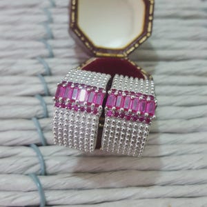 Könnte beinhalten: Ein Paar silberfarbene Ohrringe mit rechteckigem Design. Die Ohrringe haben eine Reihe von leuchtend rosa rechteckigen Steinen und kleinere rosa Steine. Die Ohrringe werden in einer offenen, weinroten Schmuckschatulle präsentiert.