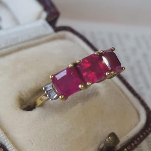 Op de afbeelding: Een gouden ring met twee vierkant geslepen, levendig rode robijnen naast elkaar, geflankeerd door kleine, heldere baguette-geslepen stenen aan de zijkant. De ring zit in een juwelendoosje met een crèmekleurige fluwelen voering.