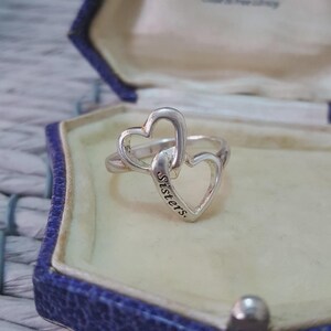 May include: A silver ring with two interlocking heart shapes. The word "Sisters" is engraved on one heart. The ring is presented in a blue jewellery box, implying a thoughtful gift.
