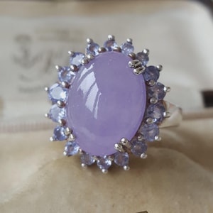 Könnte beinhalten: Ein Silberring mit einem großen, ovalen, glatten lavendelfarbenen Cabochon-Stein in der Mitte. Der zentrale Stein ist von einem Kranz aus kleineren, runden, facettierten hellblauen Steinen umgeben, die in Silberfassungen blütenartig angeordnet sind.