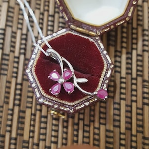 Può includere: Una collana d'argento con un ciondolo a forma di fiore con cinque pietre preziose rosa. Il ciondolo è esposto all'interno di un portagioie bordeaux con finiture dorate, su sfondo marrone.