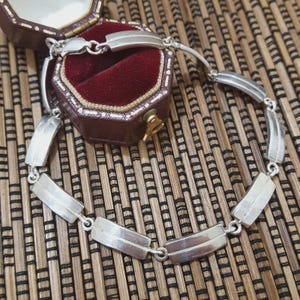 May include: A silver bracelet with rectangular and curved links, displayed near a vintage jewellery box with a burgundy velvet interior. The bracelet is on a bamboo mat.