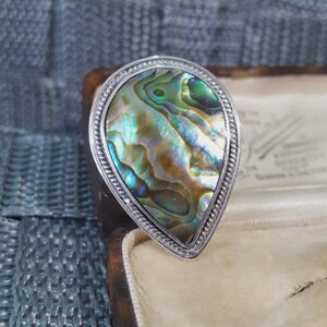 May include: A silver ring featuring a pear-shaped abalone shell. The shell has iridescent colours of green, blue and gold. The ring is set in a jewellery box. The ring's border has a rope-like design.