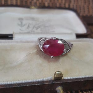 May include: A silver ring featuring a large, oval-cut ruby. The deep red ruby is the focal point, encircled by smaller, clear gemstones. The ring is presented in a vintage-style jewellery box, with a soft, cream-colored interior.