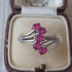 Könnte beinhalten: Ein silberfarbener Ring mit einer Ringschiene im Bypass-Stil und fünf oval geschliffenen, tief rosaroten Edelsteinen, die in einer vertikalen, leicht versetzten Reihe angeordnet sind. Die Ringschiene teilt sich in zwei glatte, polierte Stränge.
