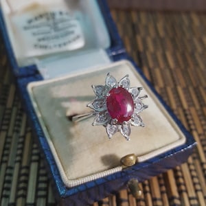 Könnte beinhalten: Ein silberner Ring mit einem großen ovalen Rubin als Mittelstein, umgeben von kleineren, klaren Edelsteinen in einem floralen Design. Der Ring wird in einer blau samtgefütterten Schmuckschatulle präsentiert.