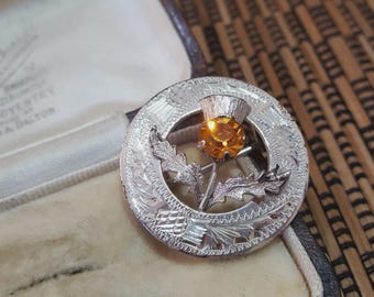 May include: A silver-toned brooch featuring a thistle design with an amber-colored gemstone center. The brooch has intricate engraved detailing around the circular frame and thistle. The brooch is displayed in a vintage jewelry box.