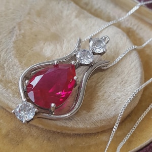 May include: A silver-toned pendant necklace featuring a central pear-shaped deep red gemstone held by four prongs. The metal frame is accented with three round clear stones, one at the base and two at the top, connected to a delicate silver chain.
