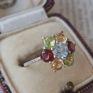 May include: A silver ring featuring a cluster of seven round faceted gemstones arranged in a floral pattern. The centre stone is light blue, surrounded by two light green, two golden yellow, and two deep red stones set in a silver band.