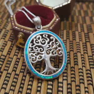 May include: Silver pendant necklace featuring an oval tree of life design with a blue and green border. The pendant is displayed in an open, brown jewelry box with a red velvet interior. The necklace chain is silver.