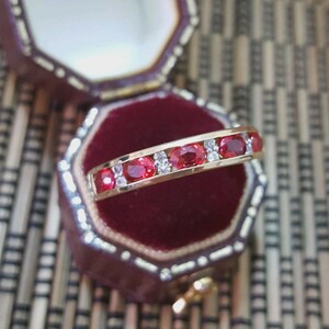 May include: A gold band ring featuring a row of alternating oval-cut red rubies and small round clear diamonds. The ring is positioned horizontally in front of an open, octagonal, dark red velvet-lined jewelry box with a gold-toned metal trim.