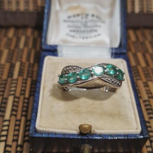 May include: A silver ring featuring five oval emeralds in a wave pattern, with small diamond accents. The ring is presented in a vintage blue jewellery box with a cream-coloured interior.