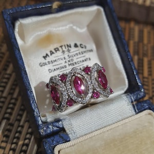 Op de afbeelding: Een zilveren ring met een centrale ovale robijn, omringd door kleinere robijnen en heldere edelstenen. De ring wordt gepresenteerd in een vintage blauwe sieradendoos met de tekst "MARTIN & CO GOLDSMITHS SILVERSMITHS AND DIAMOND MERCHANTS".