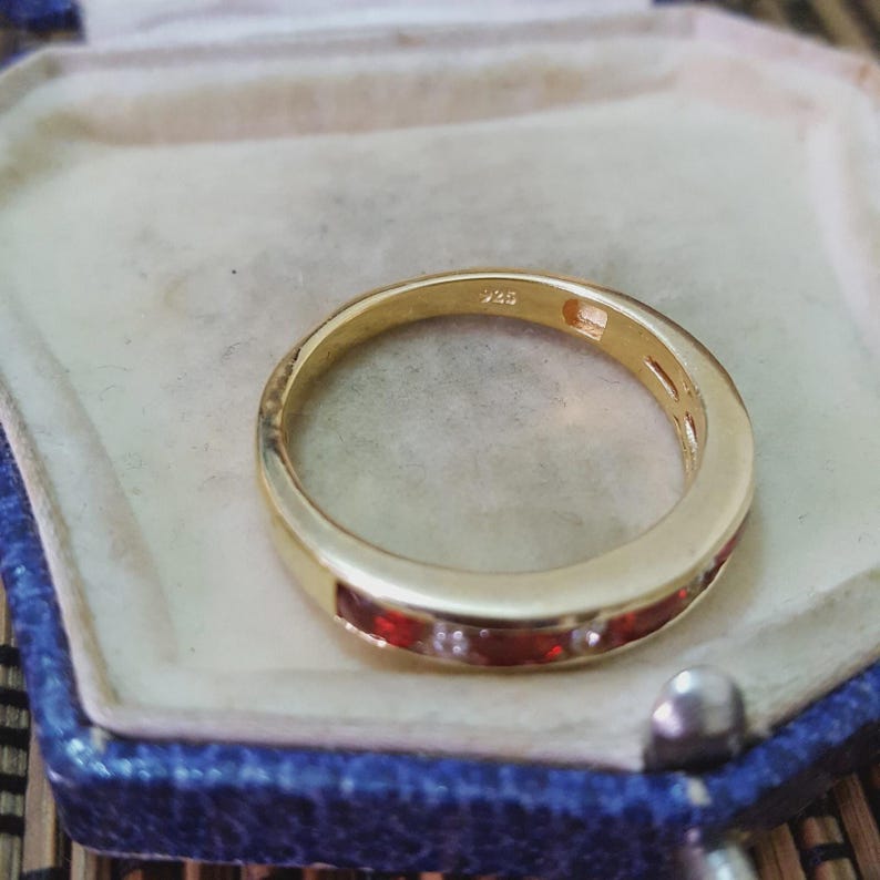 May include: A gold-toned band ring featuring a channel setting with alternating clear and deep red stones. The interior of the band is clearly stamped with the hallmark 925, indicating sterling silver construction with a gold-colored finish.