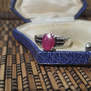 Op de afbeelding: Zilveren ring met een grote ovale robijn edelsteen in het midden, geflankeerd door kleinere zwarte stenen. De ring wordt gepresenteerd in een blauwe sieradendoos met een patroon aan de binnenkant.