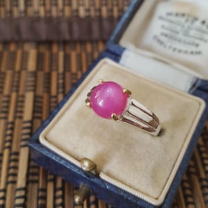Può includere: Un anello d'argento con una grande gemma rosa rotonda, con dettagli dorati. L'anello è presentato in un portagioie di velluto blu, che ne evidenzia l'elegante design e il colore vivace. La gemma è il punto focale.