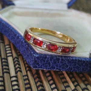 Può includere: Un anello a fascia in oro con una fila di rubini rossi taglio ovale alternati a piccoli diamanti trasparenti taglio rotondo incastonati nel canale metallico. L'anello è appoggiato sul bordo di un portagioie foderato in velluto blu.