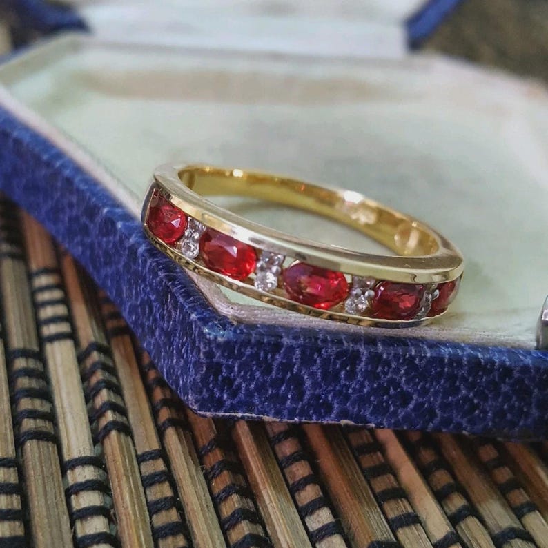 May include: A gold band ring featuring a row of alternating oval-cut red rubies and small clear round-cut diamonds set within the metal channel. The ring is displayed resting on the edge of a blue velvet-lined jewelry box.