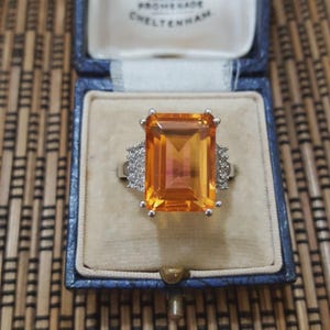May include: A silver ring featuring a large, rectangular orange gemstone. The ring is set with smaller, clear stones on either side of the main stone. Presented in a blue jewellery box with a cream-coloured interior.