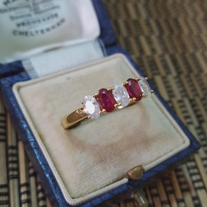 May include: A gold ring featuring a series of oval-cut red and clear gemstones. The ring is presented in a blue jewellery box with a cream-coloured interior. The gemstones are arranged in a line across the ring's top.