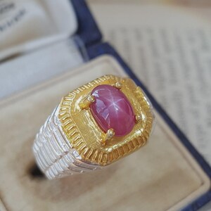 Könnte beinhalten: Ein Ring mit einem zentralen ovalen Sternrubin-Cabochon, der einen sichtbaren sechsstrahligen Sterneffekt aufweist. Der Stein ist in eine texturierte, goldfarbene Fassung eingefasst und sitzt auf einer breiten, geriffelten silberfarbenen Ringschiene.