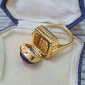 Op de afbeelding: Een gouden ring met een rechthoekige bovenkant en een scharnierend compartiment, bezet met kleine heldere stenen. De ring heeft een ronde band en een kleine, ronde, donkerblauwe steen op het scharnierende compartiment.