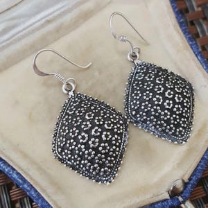 May include: A pair of silver-toned drop earrings featuring diamond-shaped pendants. Each pendant has a dark, oxidised background with a raised, textured floral pattern across the surface. The earrings are attached to simple silver-toned ear wires.